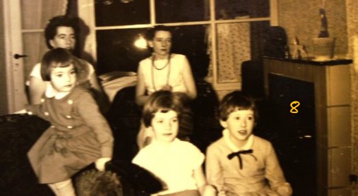 Marga en ik op de bank naast de kolenkachel. In de huiskamer zitten we tv te kijken, Marian op de leuning van de stoel, ik en Joke, Marga was net geboren en lag in de wieg.